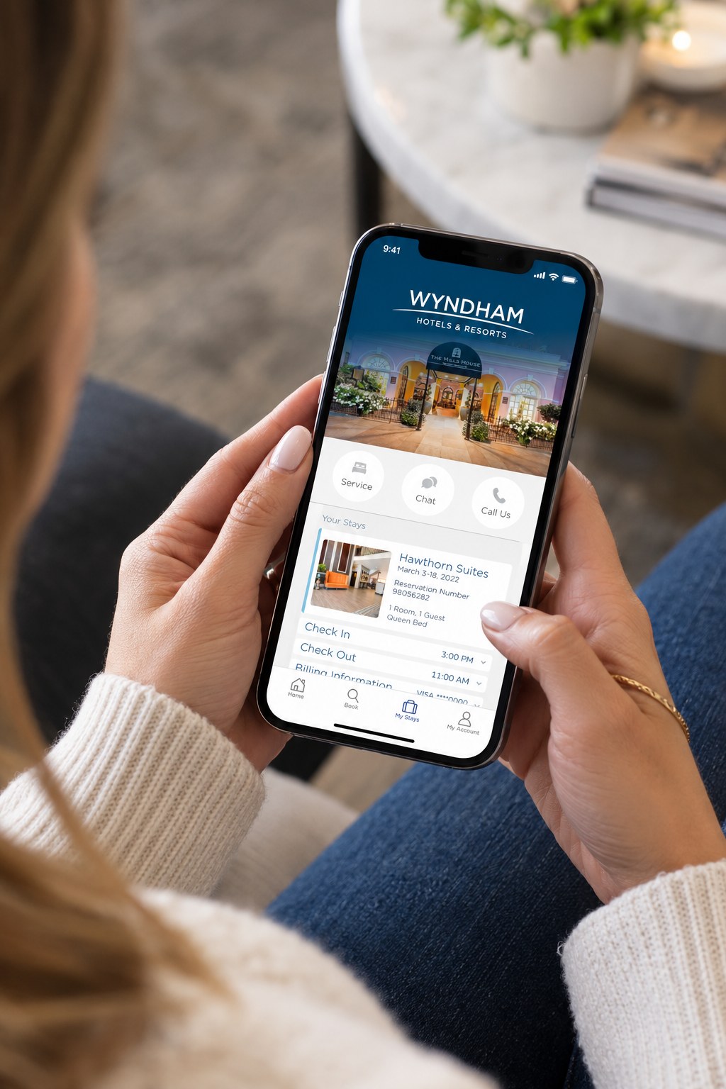 A guest holding a phone with the Wyndham app open to Your Stays: service, chat, and call options above a Hawthorn Suites reservation with check-in and check-out times.